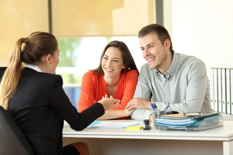 A smiling couple sits at a desk across from a businesswoman, engaged in a friendly conversation. Papers, a pen, and eyeglasses are on the desk in a bright, modern office setting.